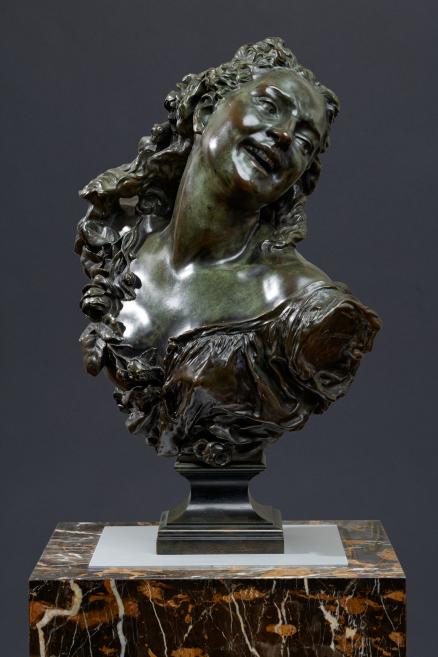 Bronze bust of a smiling woman’s head, decorated with flowers and mounted on a marble base.