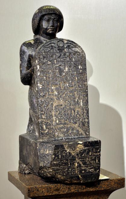 Ancient Egyptian seated stone statuette with a hieroglyphic stela on the front, displayed on a base in a museum setting.