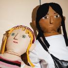 Two large textile dolls with embroidered facial features pose side by side; one has blonde hair and striped clothing, while the other has dark hair, a white shirt and a black bag.