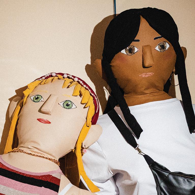 Two large textile dolls with embroidered facial features pose side by side; one has blonde hair and striped clothing, while the other has dark hair, a white shirt and a black bag.