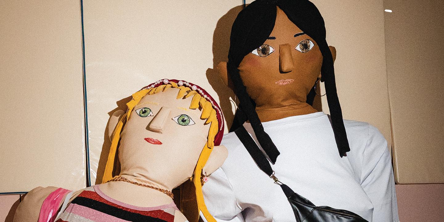 Two large textile dolls with embroidered facial features pose side by side; one has blonde hair and striped clothing, while the other has dark hair, a white shirt and a black bag.