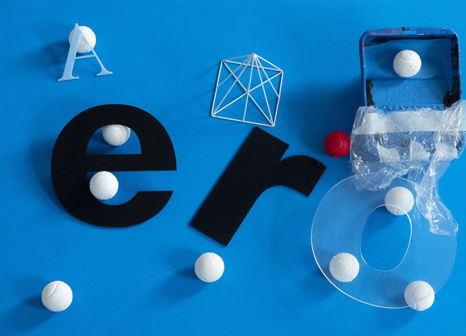 Abstract collage on a blue background with black letter forms e and r, white foam spheres, a wire geometric shape, and transparent plastic elements.