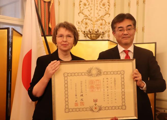 Sieviete un vīrietis tur lielu ierāmētu Japānas apbalvojuma sertifikātu, stāvot pie Japānas karoga.