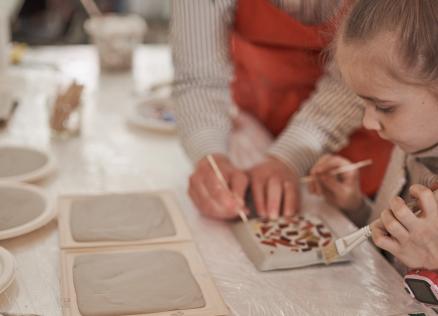 Bērns kopā ar pieaugušo dekorē keramikas flīzi ar otu, uz galda redzami māla izstrādājumi un radošie materiāli.
