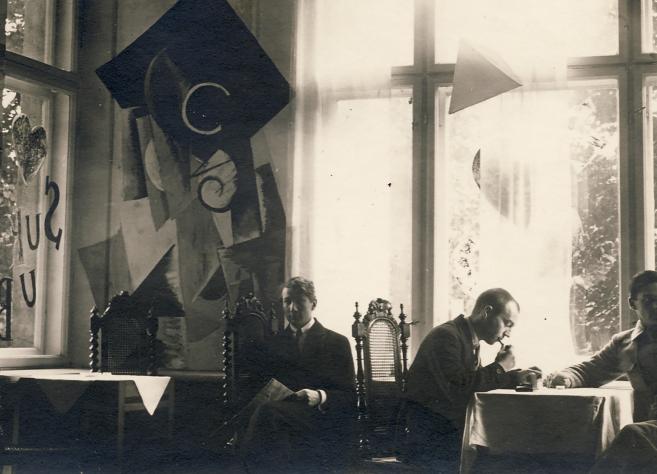 Vēsturiska melnbalta fotogrāfija ar četriem vīriešiem, kas sēž pie galda telpā ar lieliem logiem un dekoratīvām, abstraktām sienas kompozīcijām.