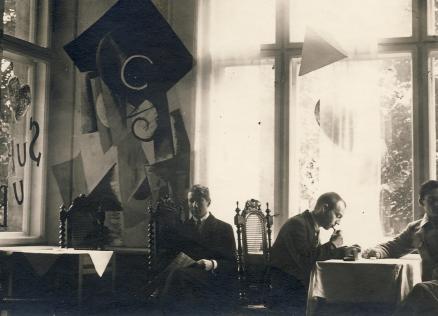 Vēsturiska melnbalta fotogrāfija ar četriem vīriešiem, kas sēž pie galda telpā ar lieliem logiem un dekoratīvām, abstraktām sienas kompozīcijām.