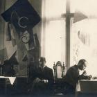 Vēsturiska melnbalta fotogrāfija ar četriem vīriešiem, kas sēž pie galda telpā ar lieliem logiem un dekoratīvām, abstraktām sienas kompozīcijām.