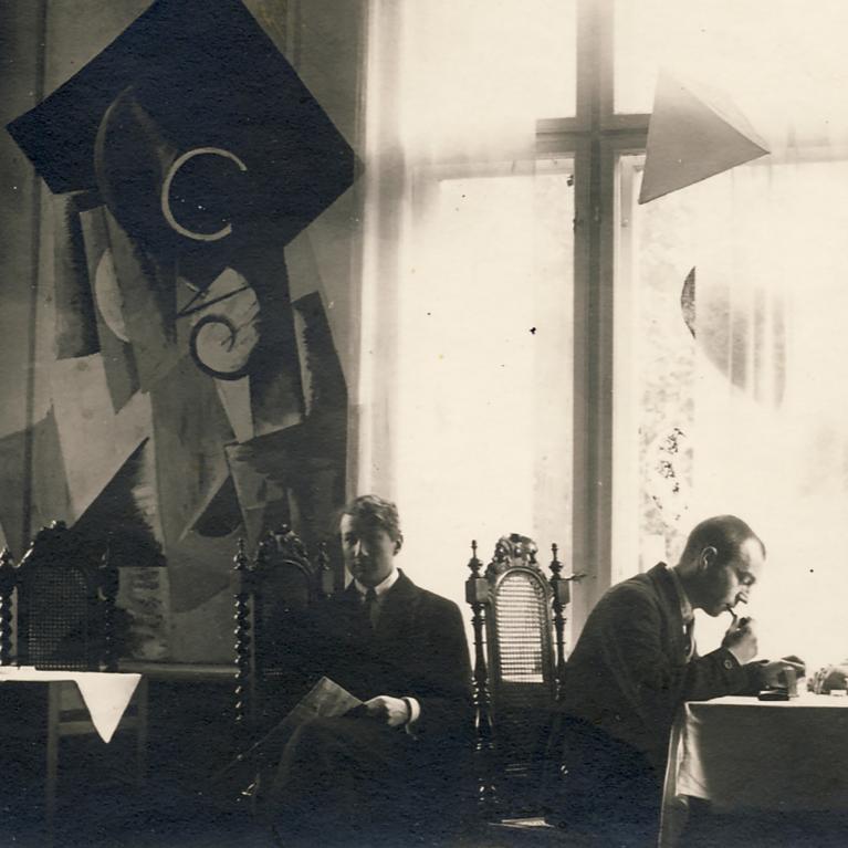 Vēsturiska melnbalta fotogrāfija ar četriem vīriešiem, kas sēž pie galda telpā ar lieliem logiem un dekoratīvām, abstraktām sienas kompozīcijām.