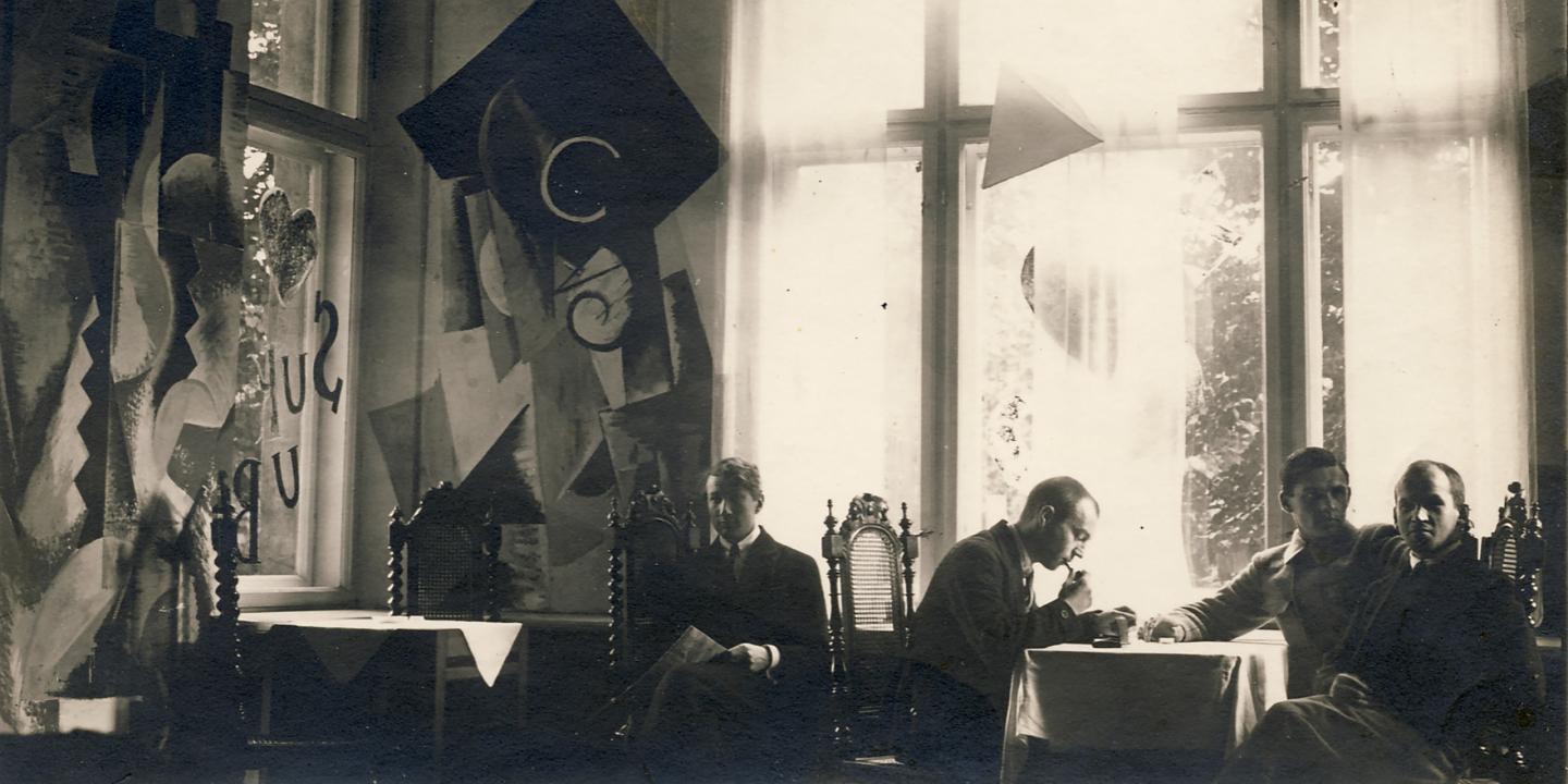 Vēsturiska melnbalta fotogrāfija ar četriem vīriešiem, kas sēž pie galda telpā ar lieliem logiem un dekoratīvām, abstraktām sienas kompozīcijām.