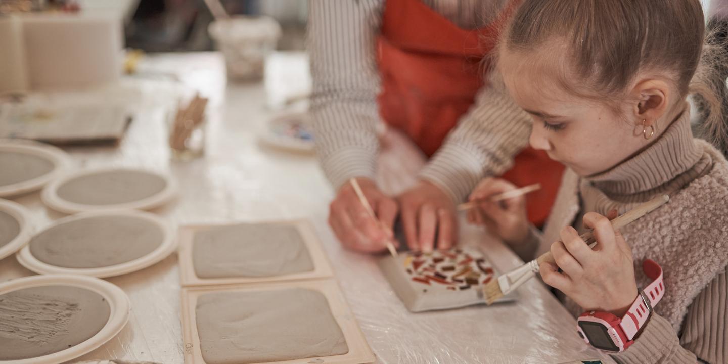 Bērns kopā ar pieaugušo dekorē keramikas flīzi ar otu, uz galda redzami māla izstrādājumi un radošie materiāli.