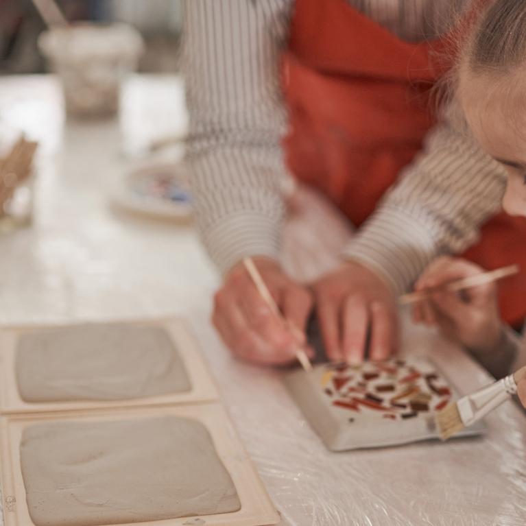 Bērns kopā ar pieaugušo dekorē keramikas flīzi ar otu, uz galda redzami māla izstrādājumi un radošie materiāli.