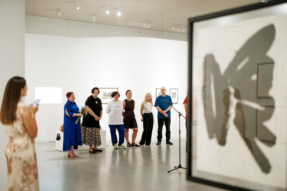Mākslas galerijā pie baltas sienas stāv seši cilvēki, bet priekšplānā redzams ierāmēts darbs ar tumšu ēnas veida formu un mikrofona statīvs.