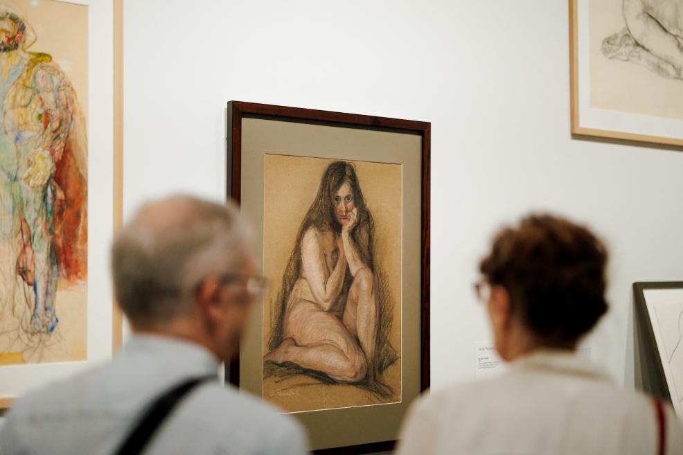 Izstāžu zālē pie baltas sienas iekārts ierāmēts sievietes akta zīmējums, ko apskata divi skatītāji priekšplānā.