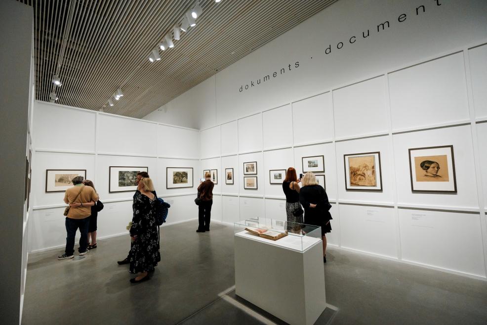 Muzeja izstāžu zāle ar pie sienām izvietotiem ierāmētiem zīmējumiem un grafikām, stikla vitrīnā centrā un apmeklētājiem, kas apskata ekspozīciju.