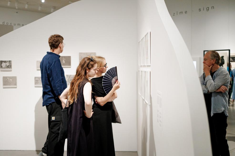 Mākslas galerijas telpā trīs apmeklētāji aplūko pie baltas sienas izvietotus darbus, bet ceturtais vīrietis stāv malā, domīgi vērojot ekspozīciju.