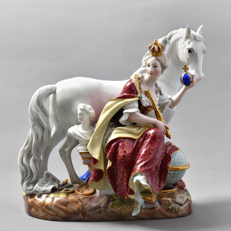 Porcelāna figūriņa ar kronētu sievieti sarkanā un zelta tērpā, kas sēž pie balta zirga un tur zilu orbītu, uz dekoratīva pamatnes.