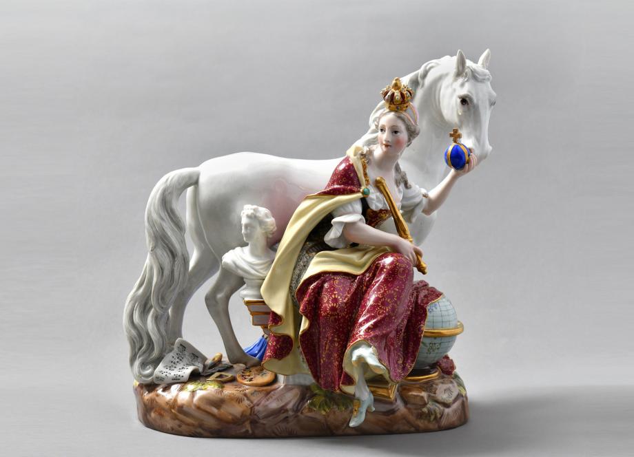 Porcelāna figūriņa ar kronētu sievieti sarkanā un zelta tērpā, kas sēž pie balta zirga un tur zilu orbītu, uz dekoratīva pamatnes.