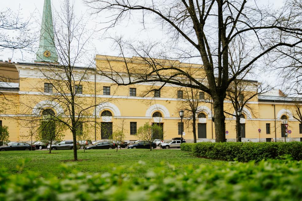 LNMM ēkas fasāde ar zaļu torni, redzama aiz koku zariem un zāliena parkā.