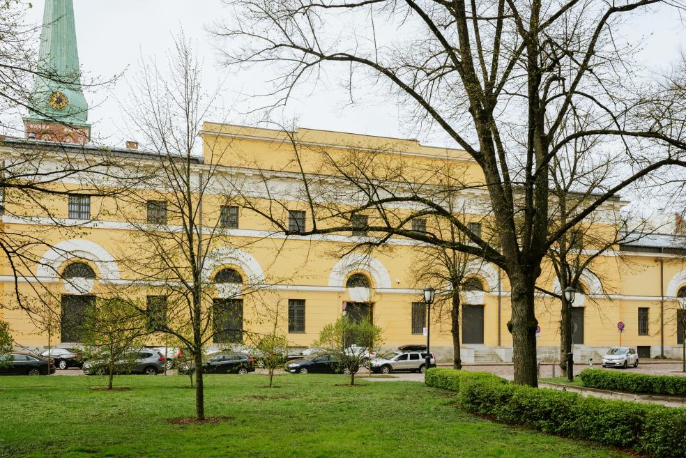 Latvijas Nacionālā mākslas muzeja ēkas fasāde ar dzeltenu sienu, zaļu torni un pulksteni, ko priekšplānā daļēji aizsedz kaili koki.
