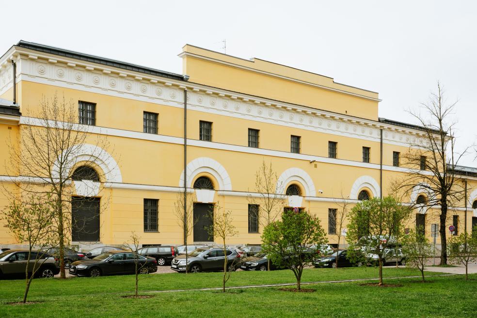 Latvijas Nacionālā mākslas muzeja dzeltenā vēsturiskā ēkas fasāde ar arkveida logiem, kokiem un stāvošām automašīnām priekšplānā.