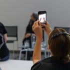 Cilvēku grupa semināra zālē, kur viens apmeklētājs ar viedtālruni filmē vai fotografē uz skatuves notiekošo.
