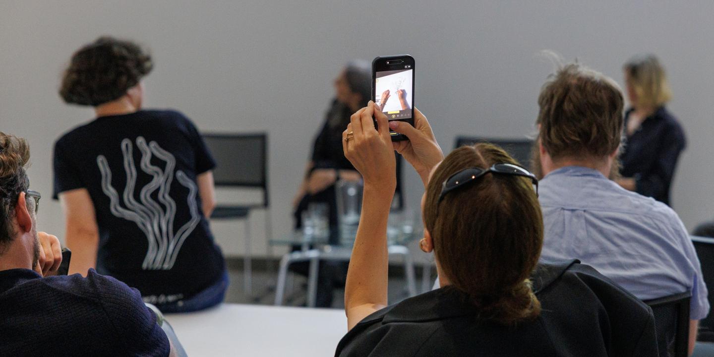 Cilvēku grupa semināra zālē, kur viens apmeklētājs ar viedtālruni filmē vai fotografē uz skatuves notiekošo.