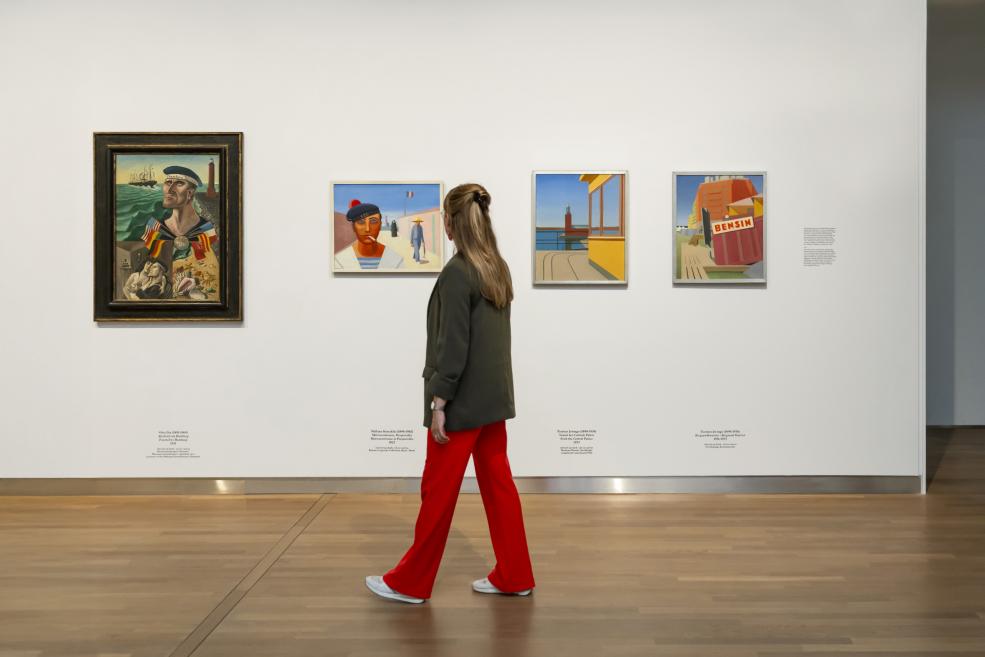 Sieviete muzeja zālē raugās uz diviem modernisma portretiem, kas izstādīti uz spilgti oranžas sienas.