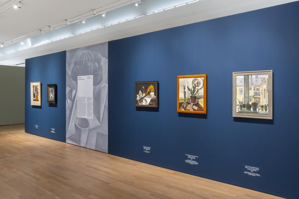 Uz tumši zilas izstāžu sienas eksponētas vairākas klusās dabas un modernisma gleznas.