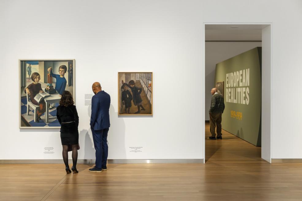 Divi cilvēki skatās gleznas uz baltas sienas, fonā redzams ieejas skats ekspozīcijas nākamajā telpā.