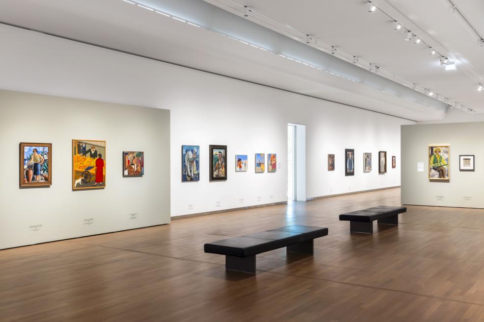 Plaša izstāžu zāle ar rindā izvietotām gleznām un diviem melniem soliņiem centrā.