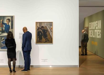 Divi izstādes apmeklētāji aplūko gleznas uz balta sienas fona, kamēr tālāk telpā citi skatītāji iepazīst ekspozīciju “European Realities 1919–1939”.
