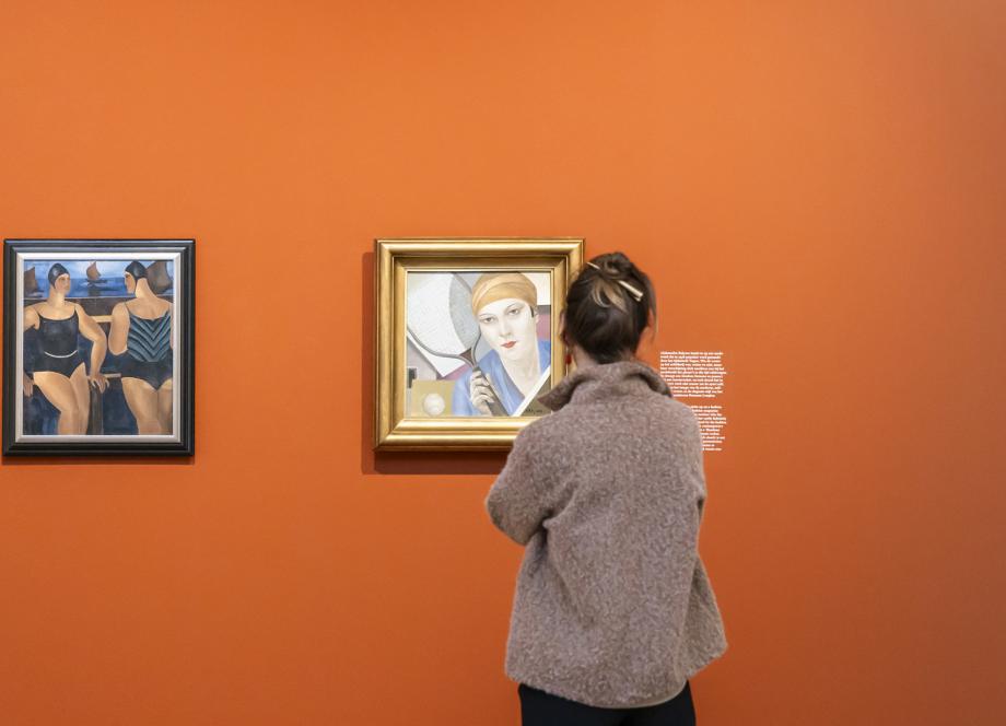 Apmeklētāja stāv pie oranžas sienas un tuvplānā aplūko divus blakus izvietotus gleznu portretus izstāžu zālē.