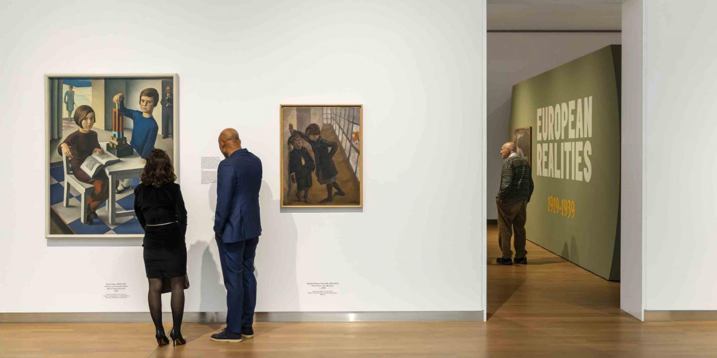 Divi izstādes apmeklētāji aplūko gleznas uz balta sienas fona, kamēr tālāk telpā citi skatītāji iepazīst ekspozīciju “European Realities 1919–1939”.