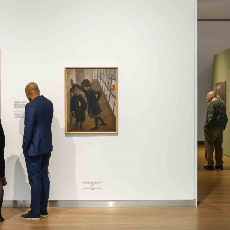 Divi izstādes apmeklētāji aplūko gleznas uz balta sienas fona, kamēr tālāk telpā citi skatītāji iepazīst ekspozīciju “European Realities 1919–1939”.