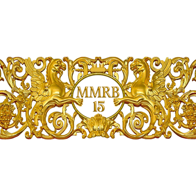 A gilded decorative ornament featuring two winged griffins and a central medallion inscribed “MMRB 15”, surrounded by scrolling floral motifs.