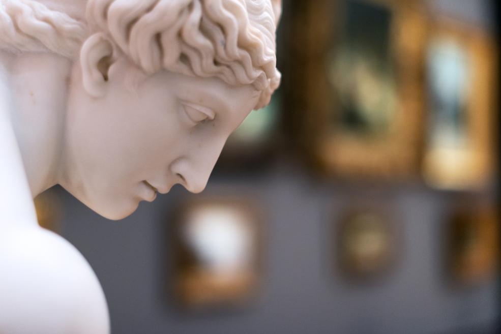 Balta marmora jaunekļa statujas profila tuvplāns ar noliektu galvu, uz neskaidra muzeja interjera fona.