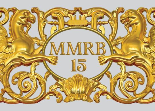 Zeltīts dekoratīvs ornaments ar diviem spārnotiem grifiem, vīnogu ķekariem un centrālu medaljonu ar uzrakstu “MMRB 15”.