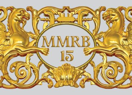 Zeltīts dekoratīvs ornaments ar diviem spārnotiem grifiem, vīnogu ķekariem un centrālu medaljonu ar uzrakstu “MMRB 15”.