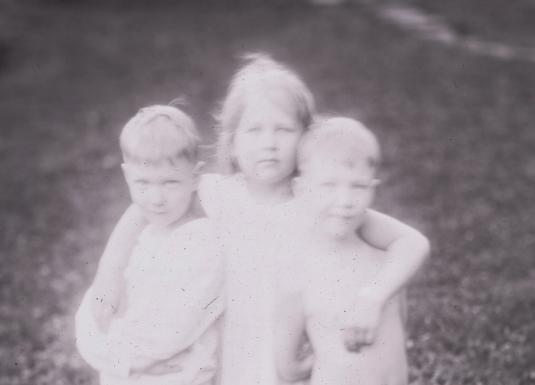Melnbalta fotogrāfija ar trīs kailiem bērniem, kas cieši apskāvušies ārā uz zāliena.