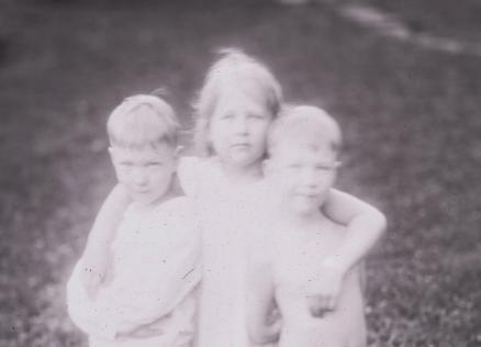 Melnbalta fotogrāfija ar trīs kailiem bērniem, kas cieši apskāvušies ārā uz zāliena.