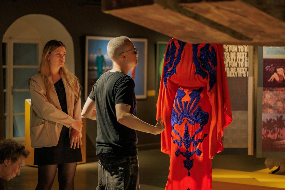 Divi apmeklētāji galerijā skatās uz koši sarkanu tērpu ar zilām izšuvuma detaļām, kas ir iekarināts izstādes telpā.