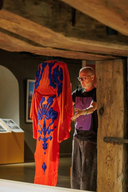 Persona muzeja zālē pie griestos piekārtas oranžas kleitas ar tumši zilām izšuvuma detaļām.