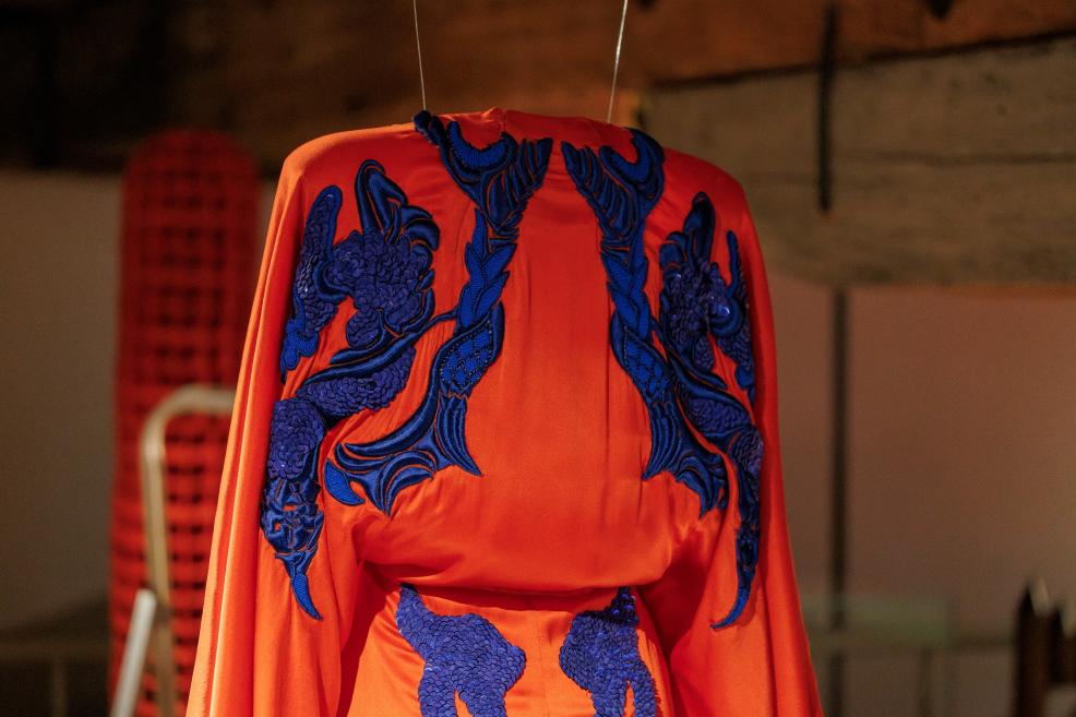 Uz pakaramā redzams spilgti oranžs tērps ar tumši ziliem izšuvumiem un reljefiem ornamentiem.