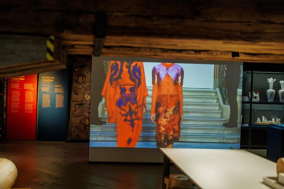 Izstāžu zālē redzama liela video projekcija ar divām modelēm spilgti oranžos un violetos tērpos, blakus informācijas stendi un vitrīnas.
