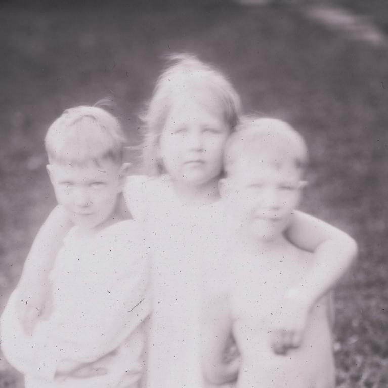 Melnbalta fotogrāfija ar trīs kailiem bērniem, kas cieši apskāvušies ārā uz zāliena.