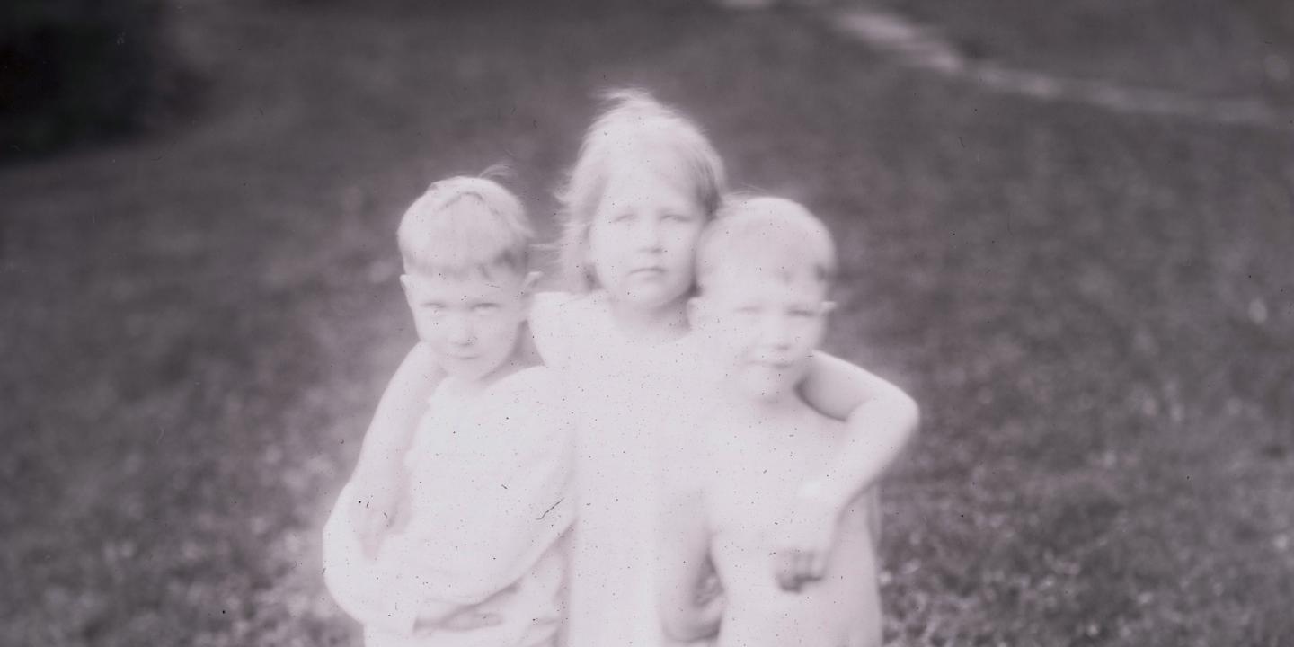Melnbalta fotogrāfija ar trīs kailiem bērniem, kas cieši apskāvušies ārā uz zāliena.