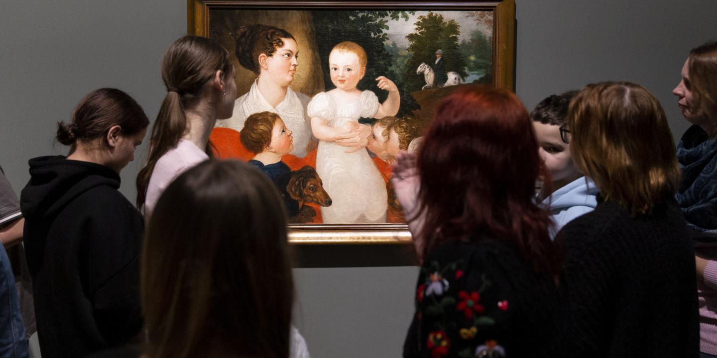 Apmeklētāju grupa muzejā stāv pie sienas un aplūko gleznu ar sievieti un bērniem.