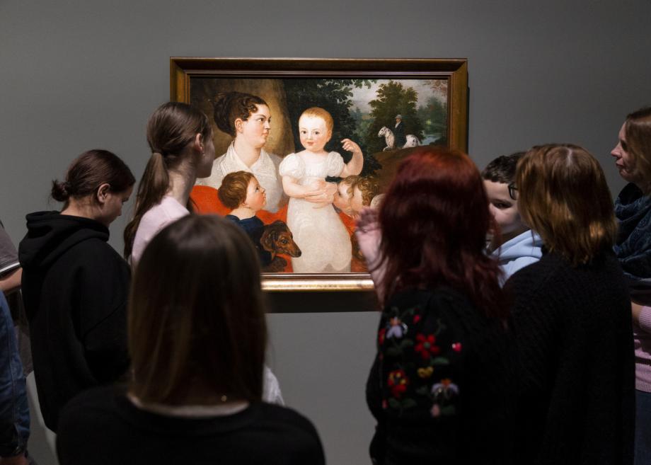 Apmeklētāju grupa muzejā stāv pie sienas un aplūko gleznu ar sievieti un bērniem.