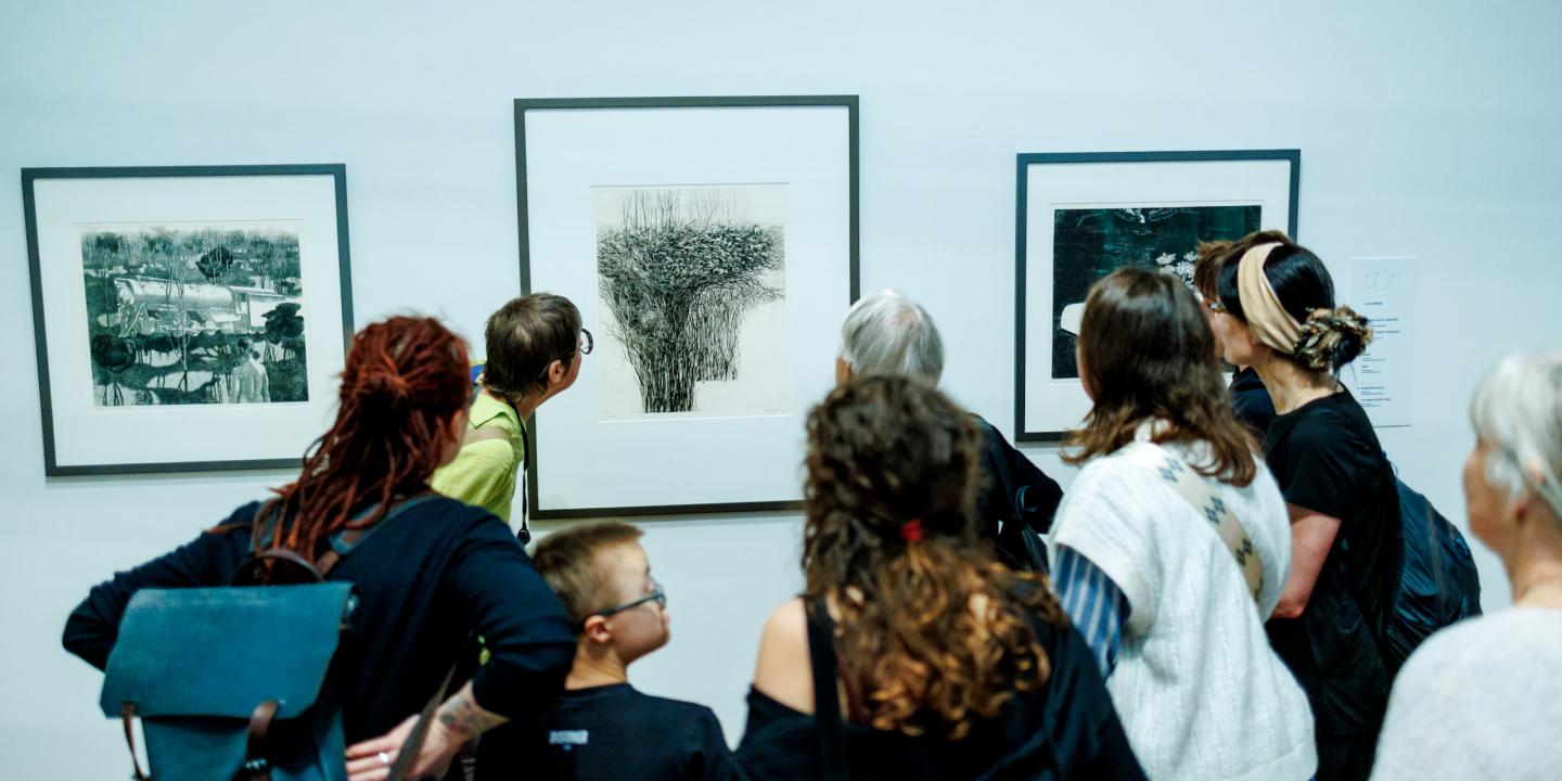 Muzeja apmeklētāju grupa stāv pie sienas un aplūko grafikas darbus rāmjos.