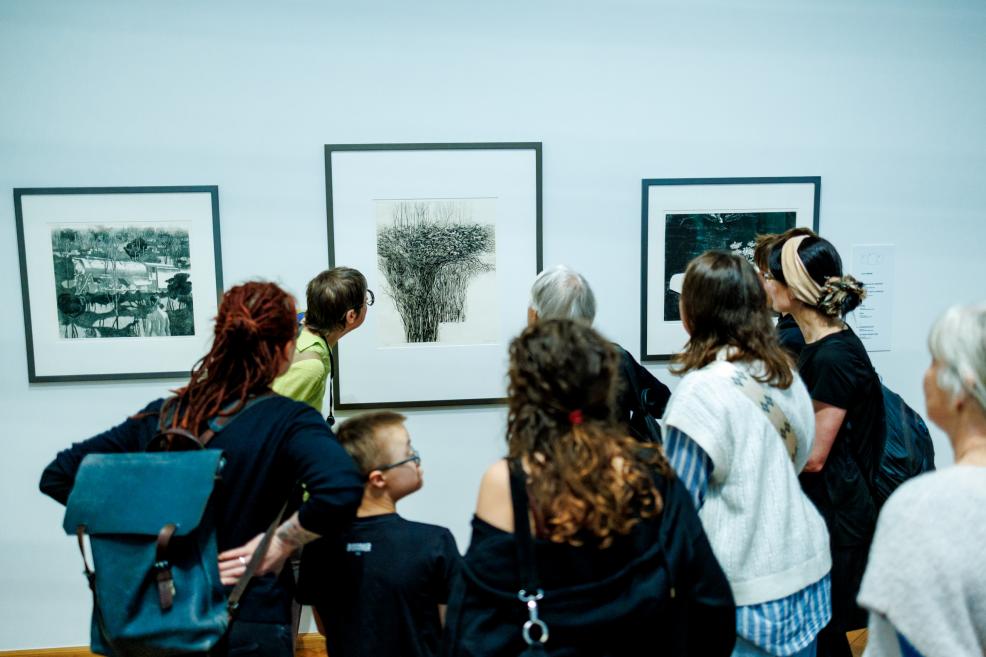 Muzeja apmeklētāju grupa stāv pie sienas un aplūko grafikas darbus rāmjos.